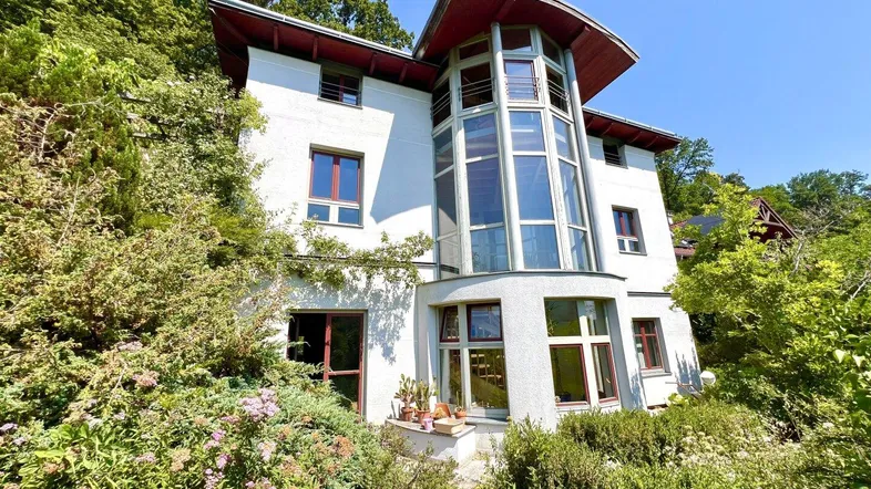 Modernes Haus mit großer Glasfassade und umgebender grüner Vegetation unter blauem Himmel.