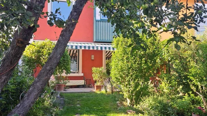 Grüner Garten mit Apfelbäumen und Blick auf die farbenfrohe Hausfassade mit Markise.