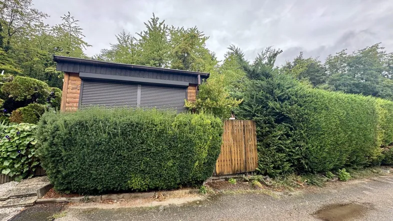 Kleines Holzhaus im Garten mit Rollläden und umgebender Hecke unter bewölktem Himmel.