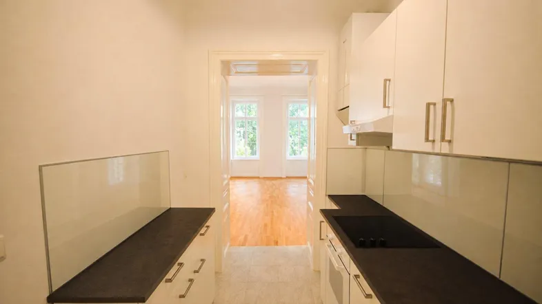 Helle Küche mit weißen Schränken, dunklen Arbeitsplatten und Blick in ein weiteres Zimmer mit Parkettboden.