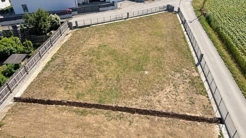 Luftaufnahme eines sonnigen Baugrundstücks mit Graben, Zaun und angrenzender Straße.