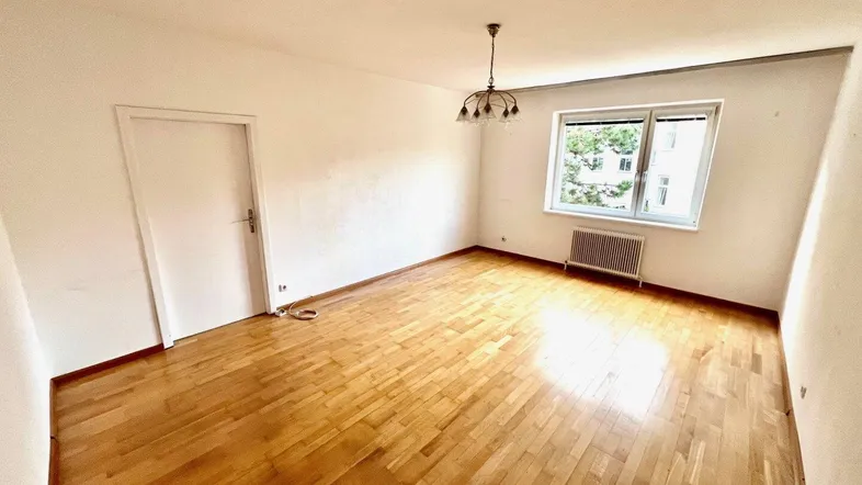 Helles, leeres Zimmer mit Parkettboden und einem Fenster, das viel Tageslicht hereinlässt.