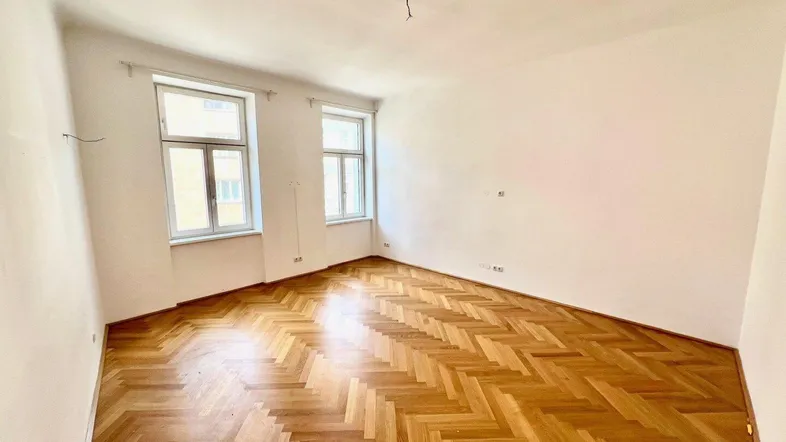 Helles Zimmer mit Fischgrätparkettboden und zwei Fenstern, bereit zur individuellen Gestaltung.