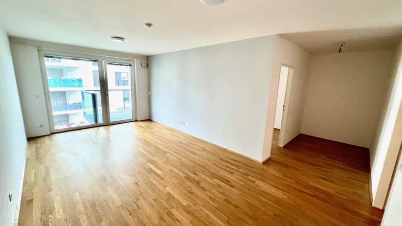 Helles, leeres Wohnzimmer mit Parkettboden und Zugang zu einem Balkon.