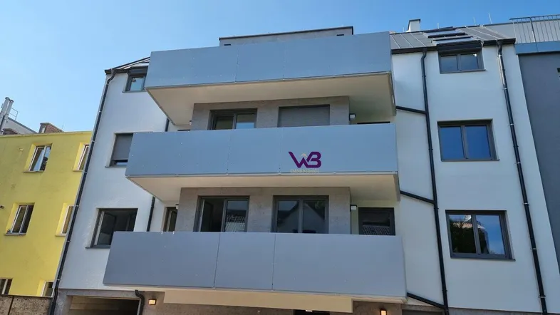 Moderne Wohnhausfassade mit mehreren Balkonen und hellgrauen Brüstungen unter blauem Himmel.