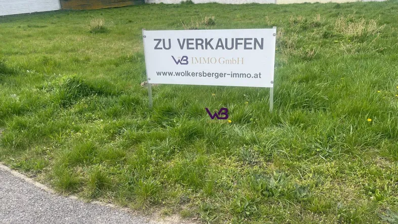 Verkaufsanzeige auf einem grünen Grundstück mit umliegenden Häusern im Hintergrund unter klarem Himmel.