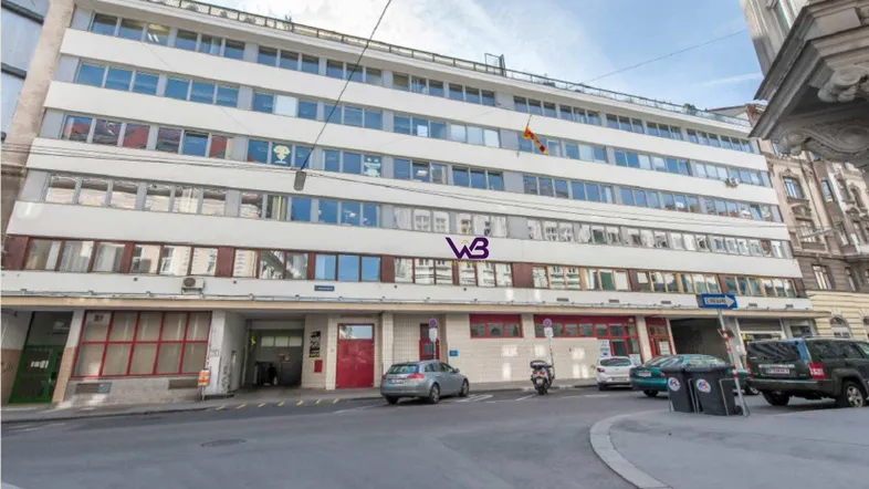 Mehrstöckiges Gebäude mit vielen Fenstern und einer Straße davor, die Parkplätze bietet.