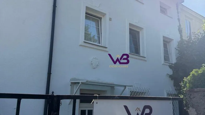 Weiß verputzte Fassade eines Mehrfamilienhauses mit einem "Zu Verkaufen"-Schild am Zaun.
