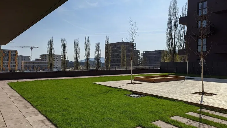 Gepflegte Grünfläche mit Holzdeck und Fliesen, umgeben von modernen Gebäuden unter blauem Himmel.