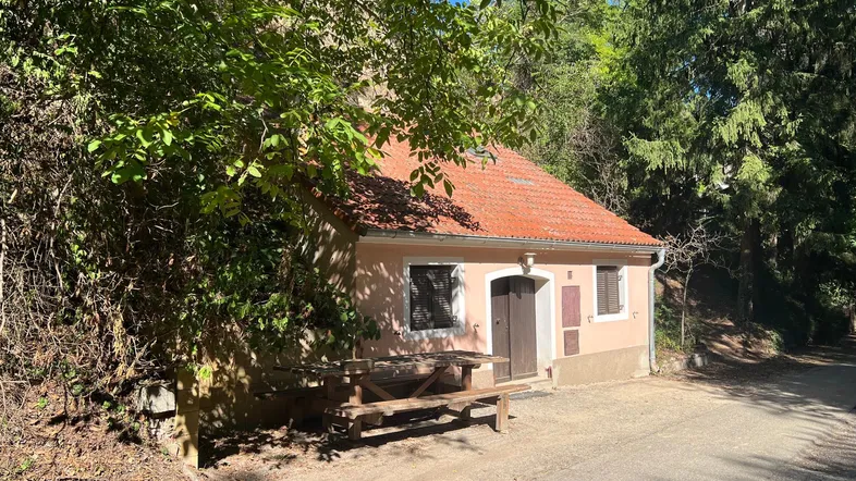 Kleines, traditionelles Haus mit rotem Ziegeldach, umgeben von Bäumen und einem Picknicktisch davor.