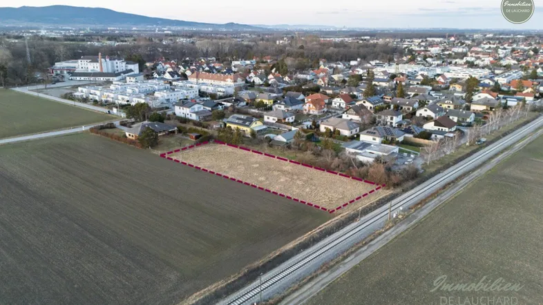 Luftaufnahme eines unbebauten Grundstücks umgeben von Feldern und Wohnbebauung mit Bergen im Hintergrund.