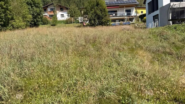 Unbebautes Grundstück mit hohem Gras und Blick auf umliegende Häuser und bewaldete Berge.