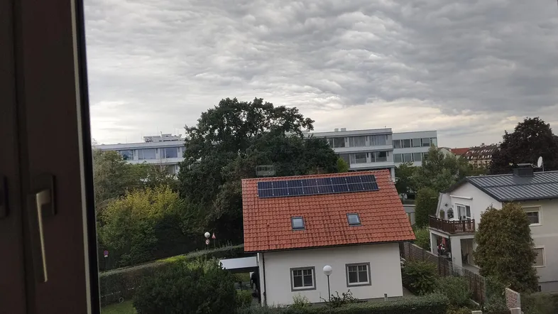 Blick aus einem Fenster auf ein Wohnhaus mit Ziegeldach und Solarpaneelen unter bewölktem Himmel.