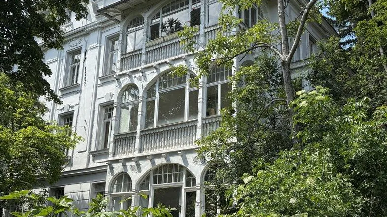 Historisches Gebäude mit eleganter Fassade und großen Fenstern, umgeben von üppigem Grün und Bäumen.