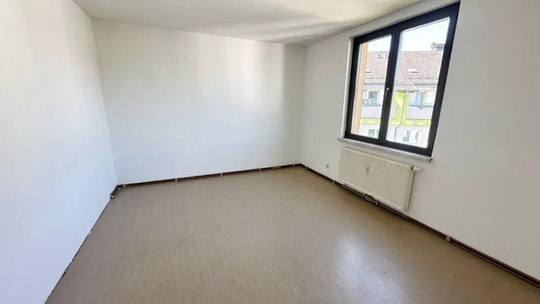 Leeres Zimmer mit weißen Wänden, Laminatboden und einem Fenster mit Heizkörper darunter.