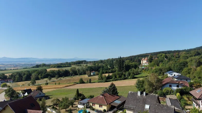 Weitläufige Luftaufnahme einer ländlichen Siedlung mit Häusern, Feldern und bewaldeten Hügeln im Hintergrund.