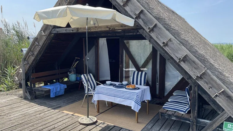 Gemütliche Terrasse mit Esstisch, Sonnenschirm und Blick auf das Wasser, umgeben von Schilf.