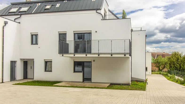 Moderne weiße Fassade eines Wohngebäudes mit dunklem Dach und Balkonen, umgeben von gepflasterten Flächen.