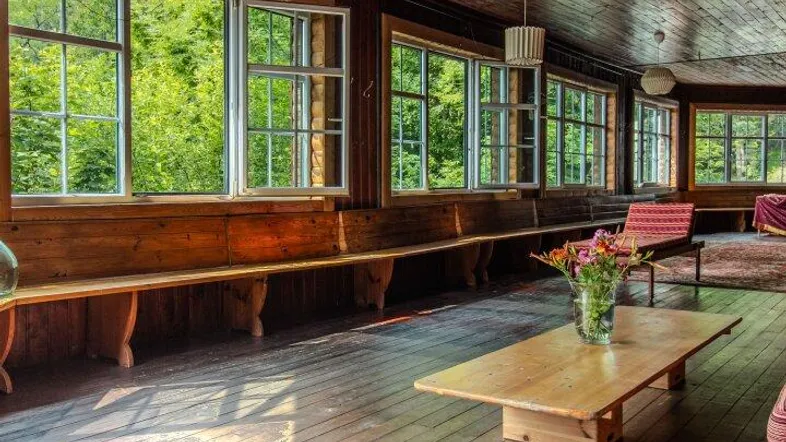 Helles, holzvertäfeltes Zimmer mit großen Fenstern und Blick ins Grüne, ausgestattet mit Holzbänken und einem Couchtisch.