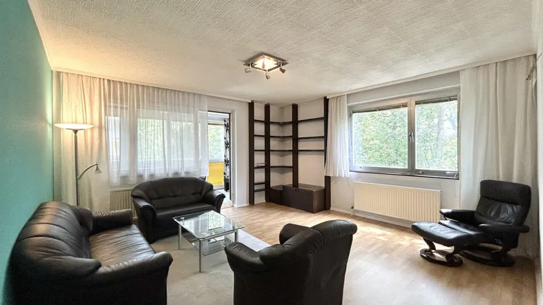 Geräumiges Wohnzimmer mit Parkettboden, bequemen Ledersofas und Zugang zum Balkon mit Blick ins Grüne.