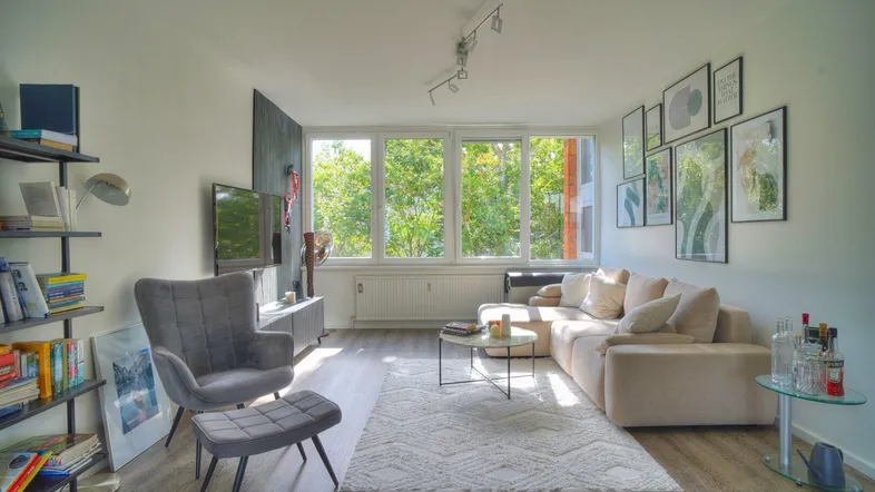 Helles Wohnzimmer mit beigem Sofa, grauem Sessel und großen Fenstern mit Blick ins Grüne.