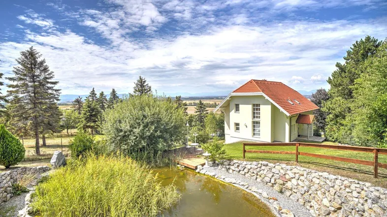 Malerisches Einfamilienhaus mit Teich im Garten und weitem Blick über die Landschaft unter blauem Himmel.