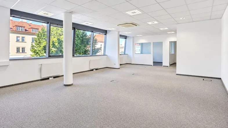 Geräumiges, leeres Büro mit Teppichboden, großen Fenstern und Blick ins Grüne.