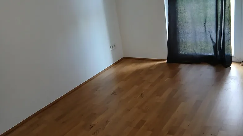Helles, leeres Zimmer mit Holzboden und großem Fenster, das viel Tageslicht hereinlässt.