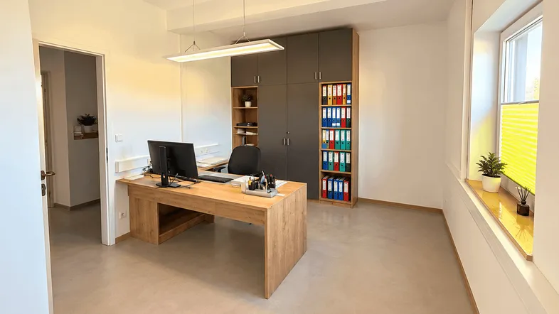 Helles und modernes Büro mit großem Schreibtisch, integrierten Schränken und viel Tageslicht durch das Fenster.