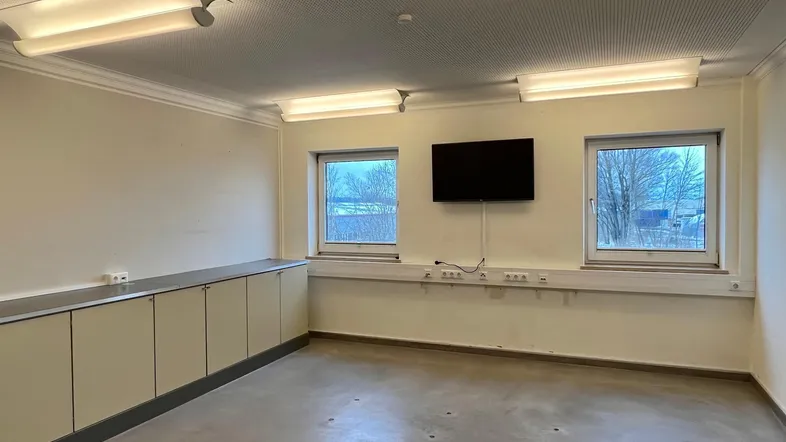 Heller Büroraum mit zwei Fenstern, Einbauschränken und einem an der Wand montierten Fernseher.