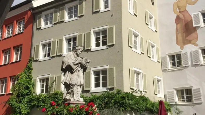 Historisches Gebäude mit einem Brunnen und einer Statue davor, umgeben von traditioneller Architektur und grünen Pflanzen.