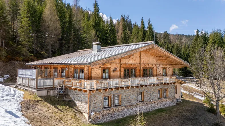 Traditionelles Holzchalet mit Steinfundament und umlaufendem Balkon, eingebettet in eine bewaldete Berglandschaft.