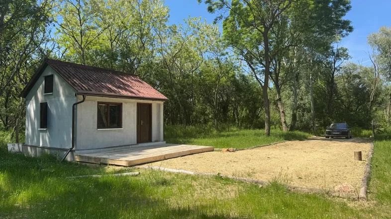 Kleines Haus mit Holzterrasse umgeben von üppiger Natur und Bäumen unter blauem Himmel.