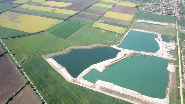 Luftaufnahme von weiten Feldern, zwei großen Wasserflächen und einem entfernten Dorf unter blauem Himmel.
