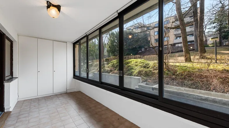 Verglaste Loggia mit Fliesenboden und Einbauschränken, bietet einen Blick auf die umliegende Grünfläche.