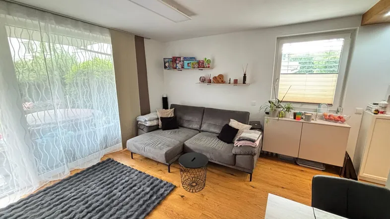Helles Wohnzimmer mit grauem Ecksofa, Holzboden und Zugang zu einem Balkon oder einer Terrasse.