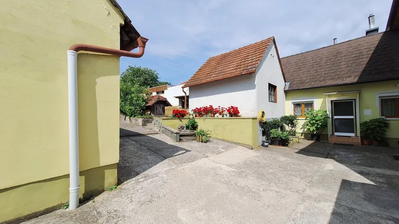 Innenhof zwischen zwei Gebäuden mit gepflastertem Bereich und Blumenbeeten unter blauem Himmel.