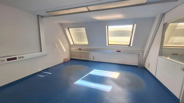 Heller Büroraum mit blauer Bodenfläche, Dachfenstern und schrägen Wänden, ausgestattet mit Heizkörper.