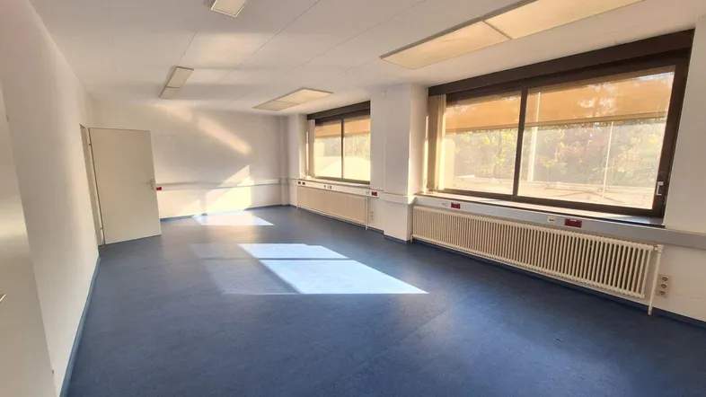 Geräumiger Büroraum mit blauer Bodenfläche, großen Fenstern und viel Tageslicht. Die Ausstattung ist funktional und gut erhalten.