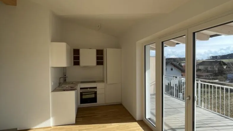 Heller offener Wohnbereich mit Holzboden, integrierter Küchenzeile und Zugang zu einem sonnigen Balkon.