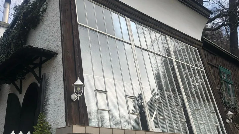 Außenansicht eines Hauses mit markanter Glasfassade und weiß verputzten Wänden unter einem bewölkten Himmel.