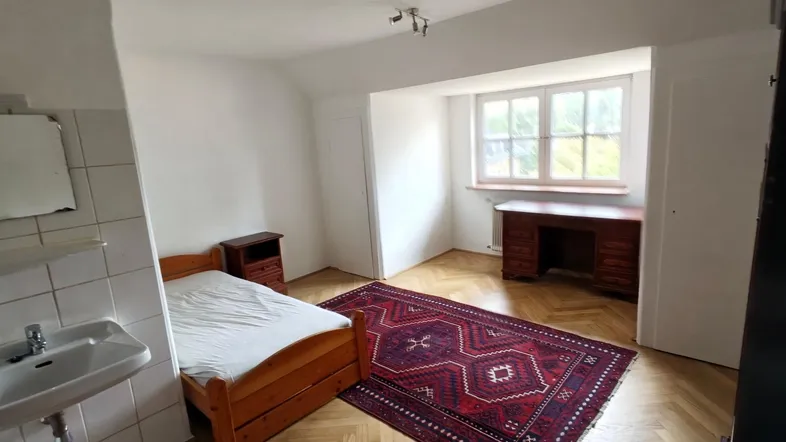 Helles Schlafzimmer mit Parkettboden, Einzelbett, Schreibtisch und integriertem Waschbecken.