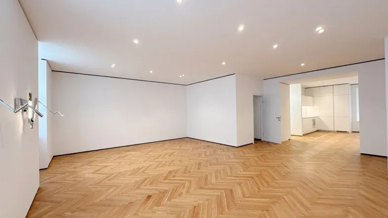Geräumiges Wohnzimmer mit elegantem Fischgrätparkett und moderner Einbauküche im Hintergrund.
