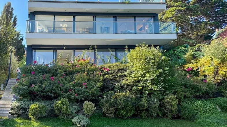 Moderne Villa mit mehreren Balkonen und einer üppigen Gartenanlage auf einem sonnigen Hanggrundstück.