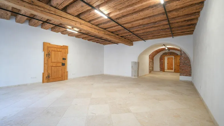 Geräumiger leerer Raum mit sichtbaren Holzbalken an der Decke, hellem Fliesenboden und einem gemauerten Bogen.