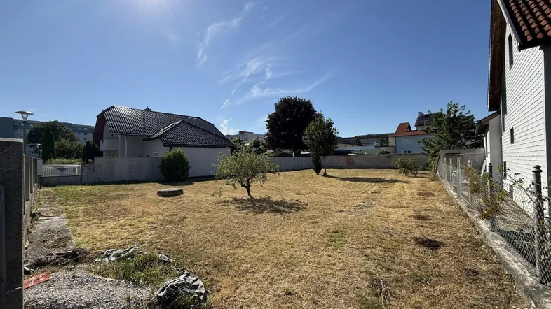 Sonniges Grundstück mit trockenem Rasen, einigen Bäumen und modernen Häusern im Hintergrund.