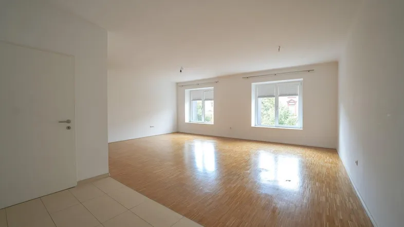 Helles, leeres Wohnzimmer mit Parkettboden und zwei Fenstern, teilweise gefliest.