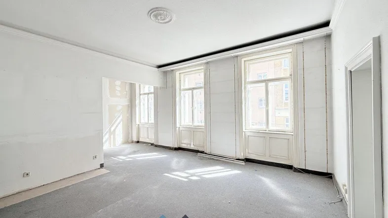 Helles Wohnzimmer im Rohzustand mit hohen Fenstern und Stuckelementen an der Decke.