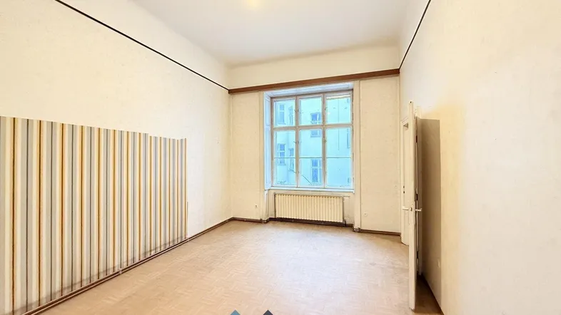 Geräumiges Zimmer mit hohen Decken, großem Fenster und Tapeten an den Wänden, bereit zur Renovierung.