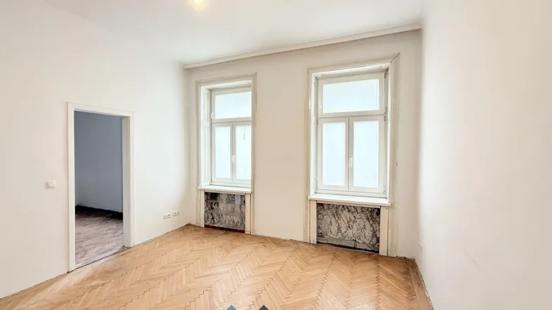 Helles, leeres Zimmer mit Fischgrätparkett und hohen Fenstern im Altbau.
